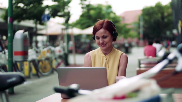 Young businesswoman with headphones and laptop sitting in cafe outdoors in city, working.