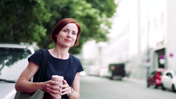 Young woman commuter with coffee standing outdoors in city, waiting for somebody.