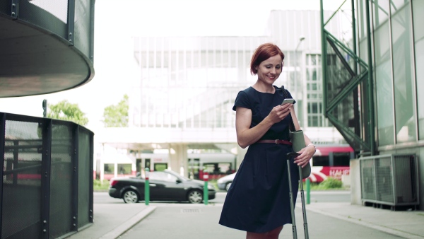 A young woman commuter with smartphone and suitcase standing outdoors in city.