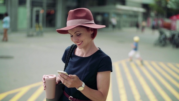 Young woman commuter with coffee and hat standing outdoors in city, using smartphone. Slow motion.