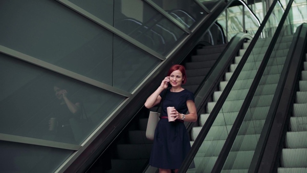 Young woman commuter with smartphone and coffee using escalator outdoors in city. Slow motion.