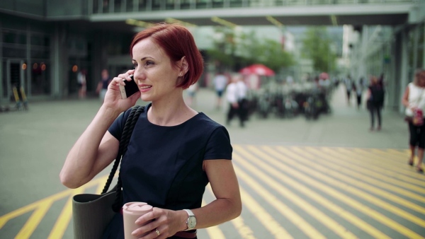 Young woman commuter with coffee and smartphone standing outdoors in city, making a call. Slow motion.