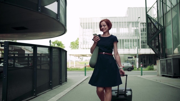 Young woman commuter with suitcase walking outdoors in city, using smartphone. Slow motion.