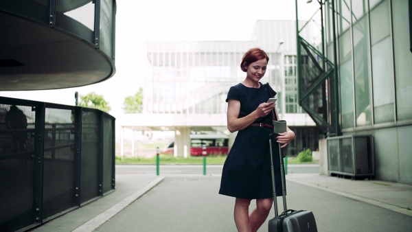 Young woman commuter with smartphone and suitcase standing outdoors in city. Slow motion.