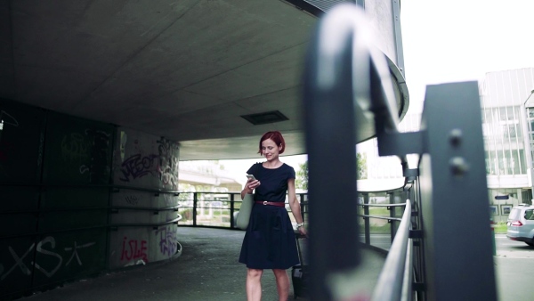 Young woman commuter with suitcase walking outdoors in city, using smartphone. Slow motion.