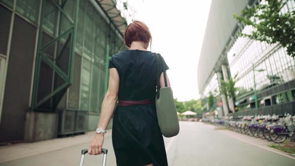 A rear view of young woman commuter with suitcase walking outdoors in city. Slow motion.