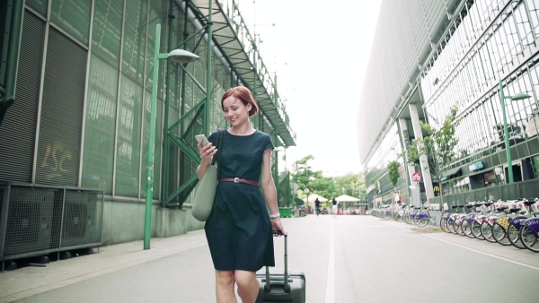 Young woman commuter with smartphone and suitcase walking outdoors in city. Slow motion.