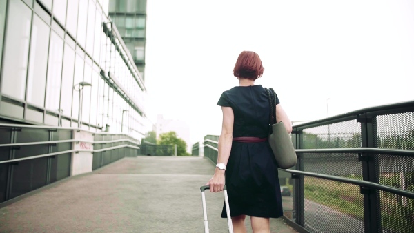 A rear view of young woman commuter with suitcase walking outdoors in city. Slow motion.