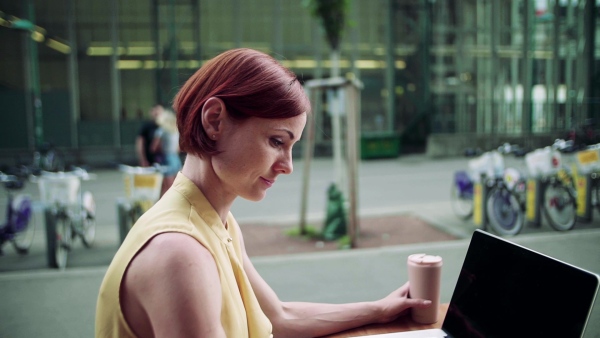 A young businesswoman with laptop sitting in cafe outdoors in city, working. Slow motion.