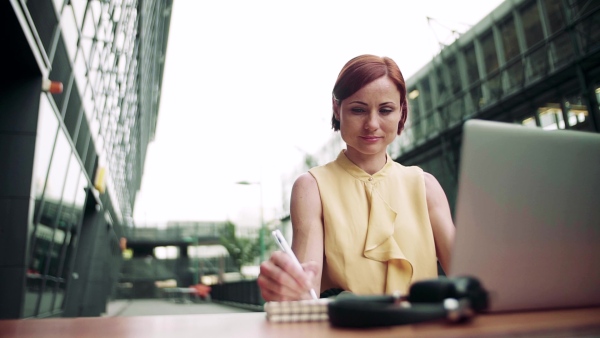 A young businesswoman with laptop sitting in cafe outdoors in city, working. Slow motion.