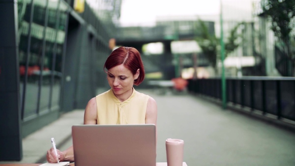 A young businesswoman with laptop sitting in cafe outdoors in city, working. Slow motion.