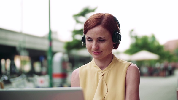 Young businesswoman with headphones and laptop sitting in cafe outdoors in city, working. Slow motion.