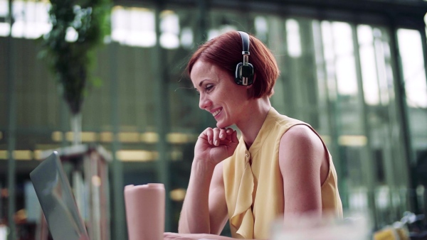 Young businesswoman with headphones and laptop sitting in cafe outdoors in city, working. Slow motion.