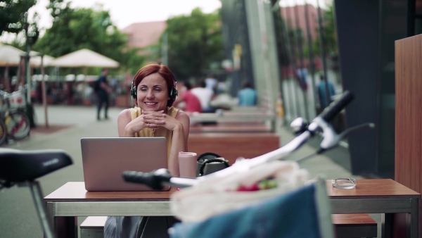 Young businesswoman with headphones and laptop sitting in cafe outdoors in city, working. Slow motion.