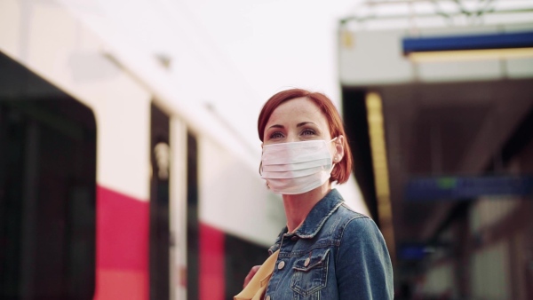 Young woman commuter with face mask standing outdoors on station in city, waiting for the train. Slow motion.