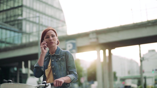 Young woman commuter with smartphone and bicycle standing outdoors in city. Slow motion.