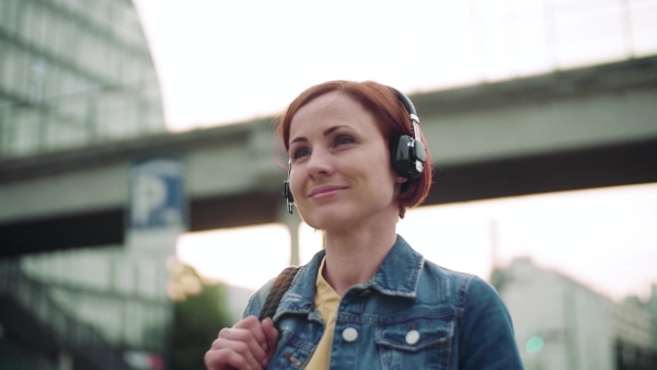 Young woman commuter standing outdoors on station in city, listening to music. Slow motion.