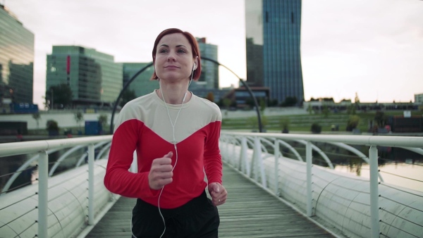 A young woman with earphones doing exercise on bridge outdoors in city, running. Slow motion.