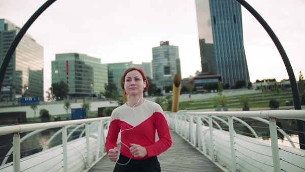 A young woman with earphones doing exercise on bridge outdoors in city, running. Slow motion.