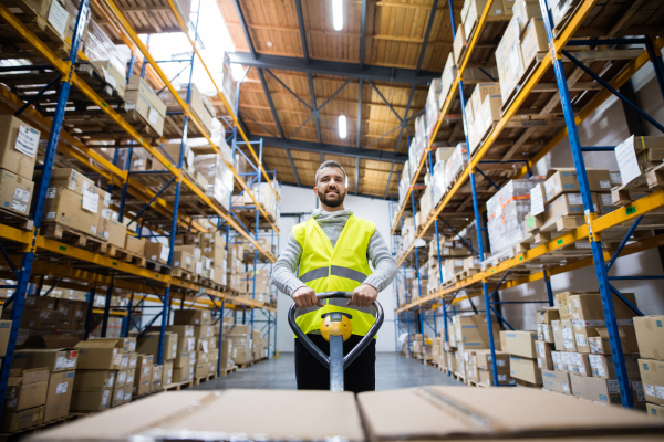 Young male warehouse worker pulling a pallet truck with boxes.