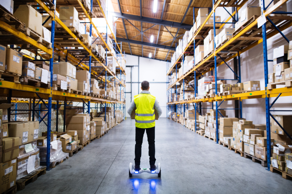 Male warehouse worker on hoverboard, holding a large box. Rear view.