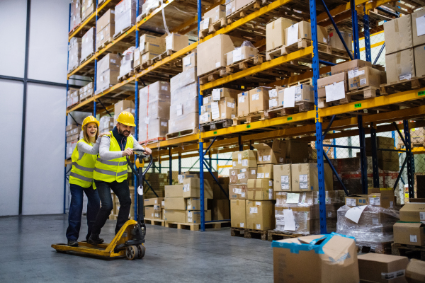 Two young workers in a warehouse, having a ride on a pallet truck.