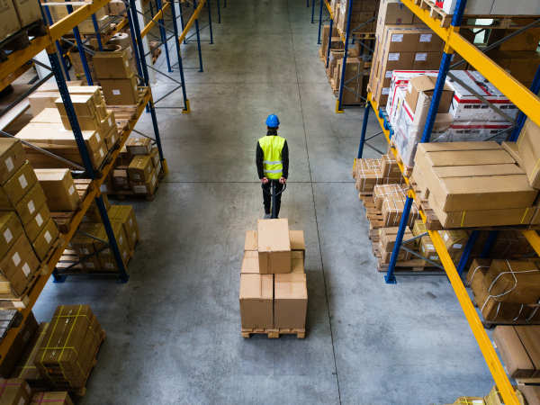 Young male warehouse worker pulling a pallet truck with boxes. Rear and high angle view.