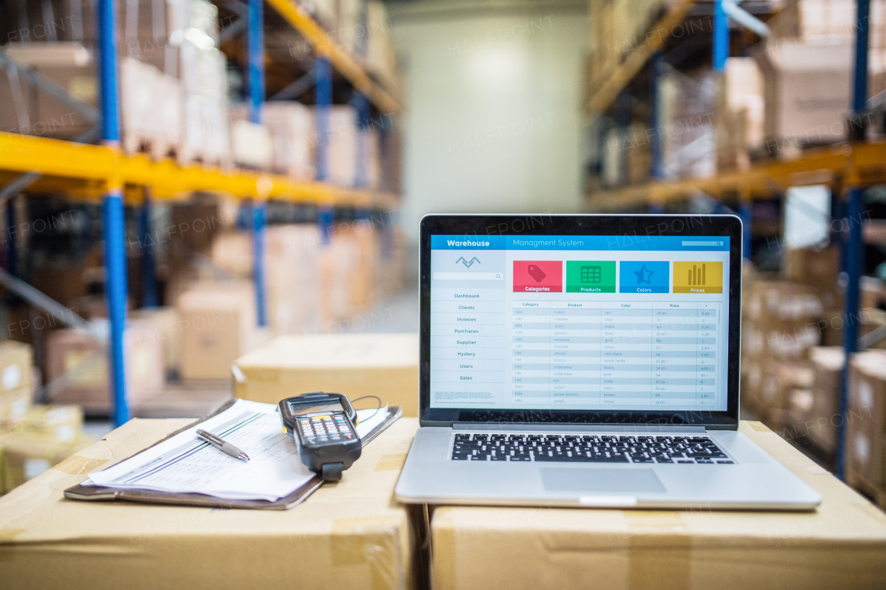 Laptop, barcode scanner, clipboard with notes and pen on the top of boxes in a warehouse.