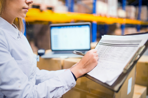 Unrecognizable young woman warehouse worker or supervisor with clipboard, controlling stock.