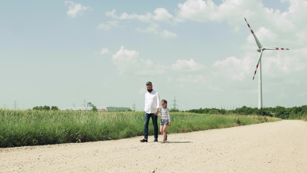 A mature father with small daughter on field on wind farm, walking.