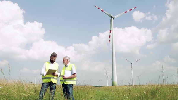 Two engineers or technicians with clipboard standing on wind farm, making notes.