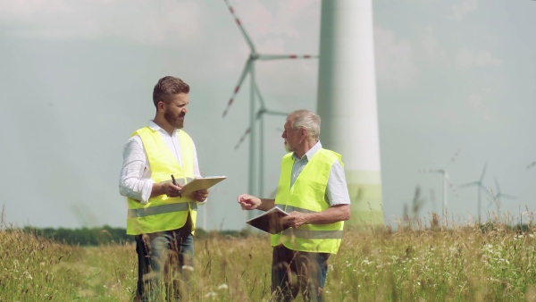 Two engineers or technicians with clipboard standing on wind farm, making notes.