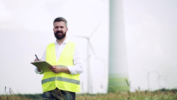 An engineer with clipboad standing on a field on wind farm, making notes.