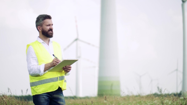 An engineer with clipboad standing on a field on wind farm, making notes.