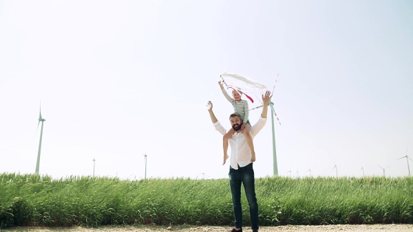 Mature father with small daughter standing on field on wind farm, giving her piggyback ride. Slow motion.