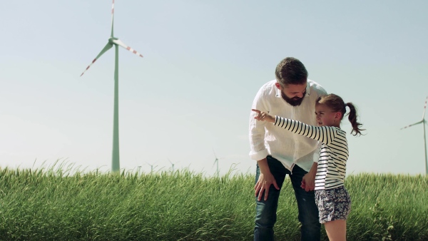 A mature father with small daughter on field on wind farm, talking.