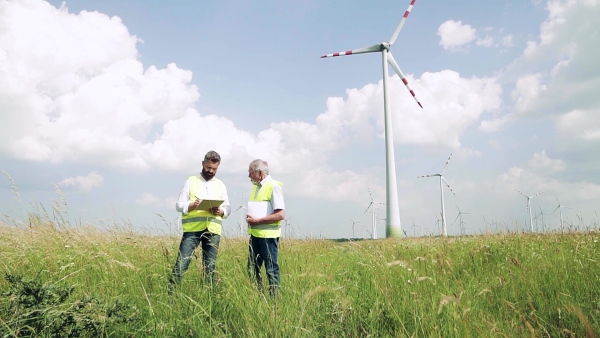 Two engineers or technicians with clipboard standing on wind farm, making notes. Slow motion.