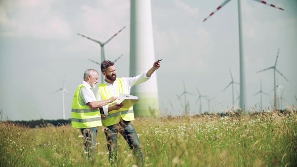 Two engineers or technicians with clipboard standing on wind farm, making notes. Slow motion.