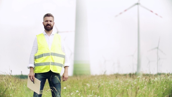 An engineer standing on a field on wind farm, looking at camera. Slow motion.