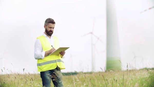 An engineer with clipboad standing on a field on wind farm, making notes. Slow motion.