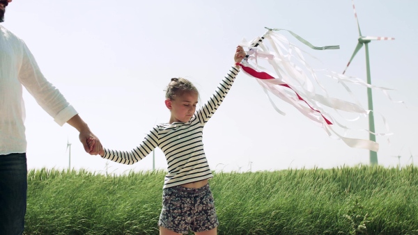 An unrecognizable father with small daughter on field on wind farm, playing with hand kite.