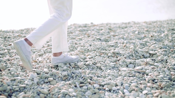 Midsection of legs of senior woman with white trousers walking on rocky beach by lake. Slow motion.