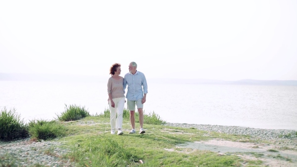 Front view of happy senior couple on a holiday walking by the lake, talking. Slow motion.