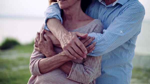 A midsection of senior couple on a holiday standing by the lake, hugging. Slow motion.