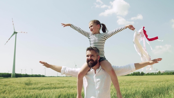 Mature father with small daughter standing on field on wind farm, giving her piggyback ride.