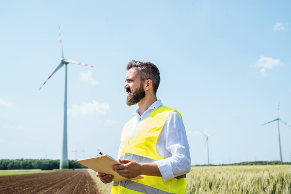 An engineer or technician with clipboard standing on a field on wind farm, making notes.