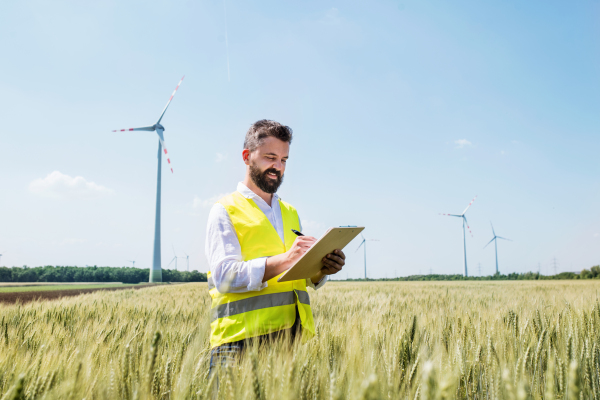 An engineer or technician with clipboard standing on a field on wind farm, making notes.