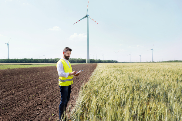 An engineer or technician with clipboard standing on a field on wind farm, making notes.