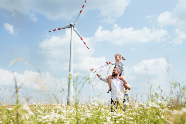 Mature father with small daughter walking on field on wind farm, giving her piggyback ride.