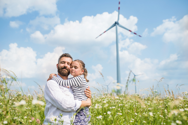 Mature father with small daughter standing on field on wind farm, eyes closed.